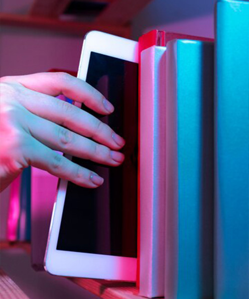 A person’s hand pulls a tablet from a row of books on a bookshelf.